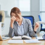 A stressed professional woman in a Singapore office removing her glasses and pinching the bridge of her nose, showing signs of workplace burnout and mental exhaustion