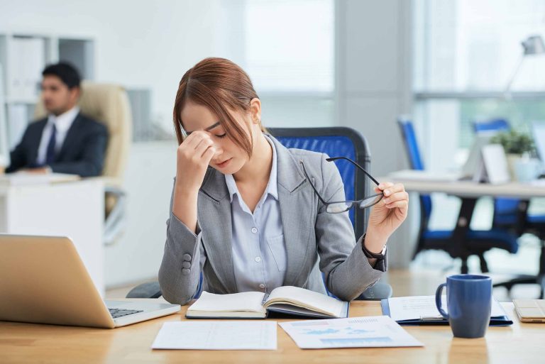 A stressed professional woman in a Singapore office removing her glasses and pinching the bridge of her nose, showing signs of workplace burnout and mental exhaustion